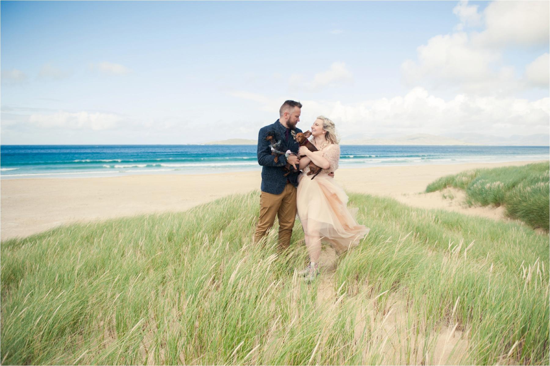 A bride and groom their their dogs stand on sand dunes on Scarista Beach for couple photos. 