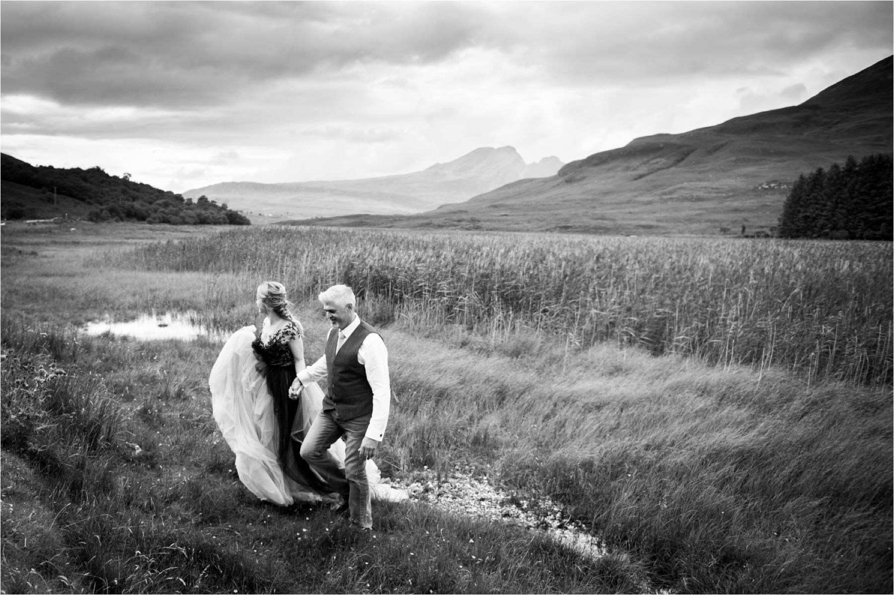 A black and white photo of a bride and groom walking across a field with storm clouds overhead. 