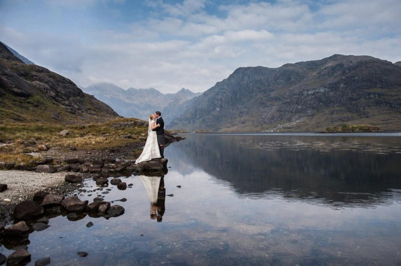 An elopement on the Isle of Skye on Loch Coruisk. A couple stands on the edge of the loch, with mountains behind them, reflected in the loch.
