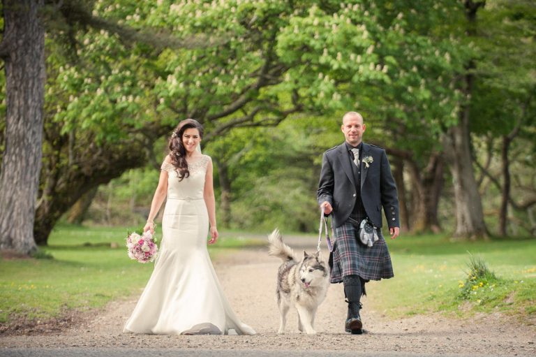 A bride, groom in a kilt and their dog walk through the grounds of Lews Castle on their wedding day.