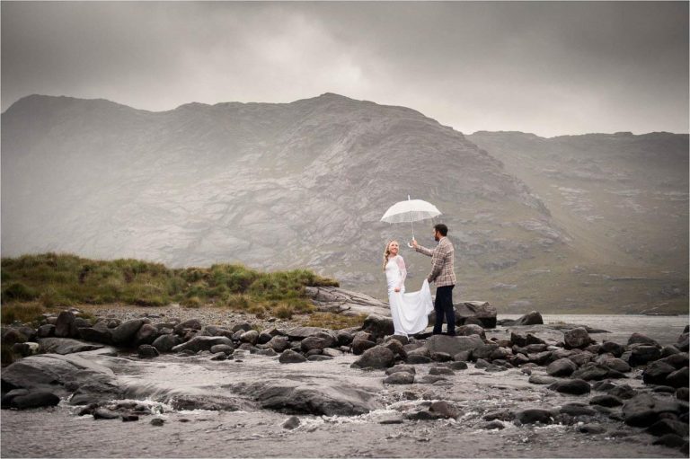 A bride and groom hold an umbrella over themselves as they walk near a river on the Isle of Skye in Scotland.