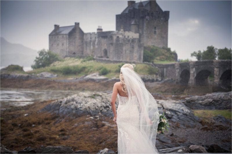 A bride in a white dress and veil walks towards Isle of Skye wedding venue Eileen Donan Castle.
